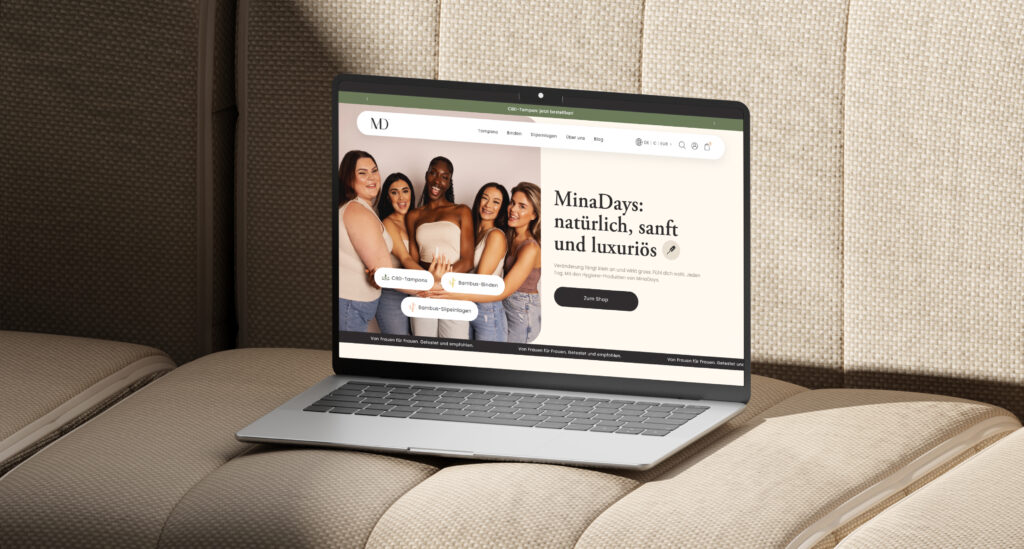 Laptop auf hellem Sofa zeigt Website mit fünf lächelnden Frauen und Text zu MinaDays Hygieneprodukten